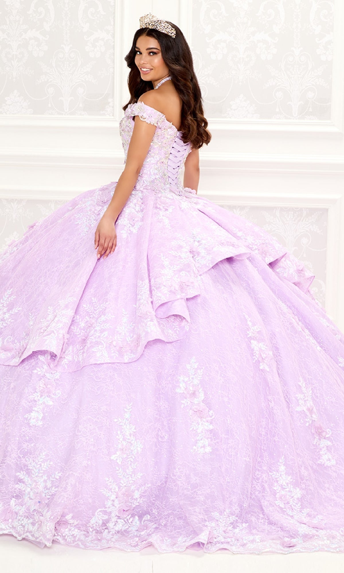 A young woman stands sideways in a white, decorative room, wearing the Quinceanera Dress PR30089 By Princesa—an ornate lavender gown with embroidered lace and a tiara.